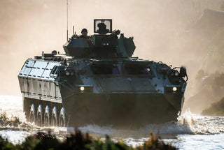 A dark green armored military vehicle with a boxy, rugged design and eight large wheels is driving through shallow water.